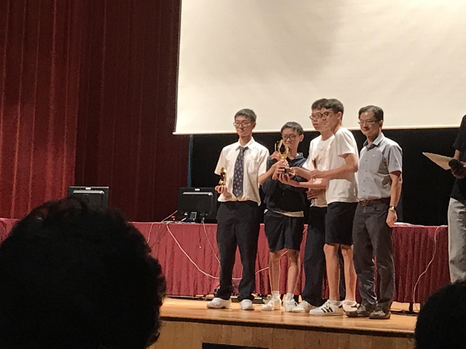 A group of students receives awards on a stage during a prize presentation ceremony, with an adult presenter handing over a trophy.